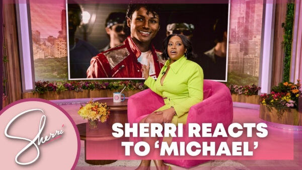 Sherri seated pointed at photo still from "Michael" movie in video wall behind her